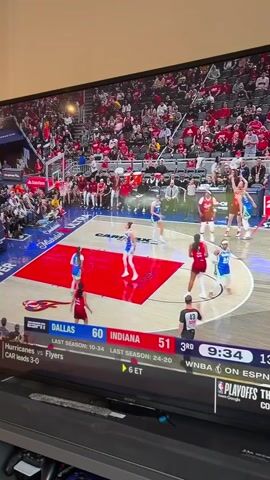 The video shows a basketball game being played on television. The teams are the Dallas Wings and the Indiana Fever. The score is 60-51 in favor of Dallas. It is the third quarter, with 9:31 left in the period. The game is being broadcast on ESPN. There are many spectators in the stands, most of whom are wearing red. The game is being played on a court that has a red section in the middle. A fan is heard yelling “yes!” while watching the game.