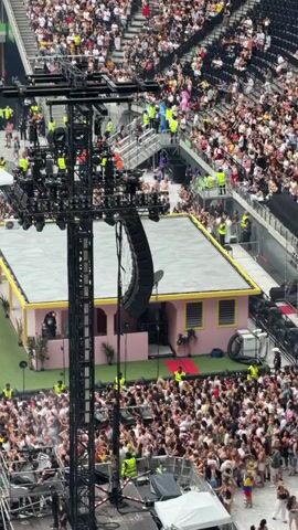 The video begins with an aerial view of a crowded stadium, focusing on a large stage setup with lighting rigs and speakers. A pink structure resembling a small house is positioned on the stage. The camera then pans across the massive audience filling the stadium seats and the area in front of the stage. A large screen displays a scene of a man and a woman sitting on a park bench, interacting. The video captures the scale of the event, with thousands of people gathered to watch a performance.