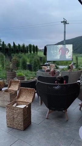 This video captures an outdoor cinema experience set in a mountainous region. The scene is viewed from a slightly elevated perspective, looking towards a large projection screen displaying a film. Several people, mostly seen from behind, are seated in woven outdoor chairs, many wearing headphones, indicating an audio component to the viewing. The environment suggests a casual, relaxed setting, with plants in pots scattered around and a rustic, woven storage bin in the foreground. The backdrop features lush green mountains and a cloudy sky, with a ski lift tower visible in the distance. The overall impression is one of people enjoying a movie under the open sky amidst natural scenery.