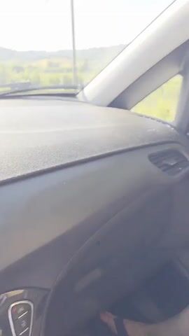 The video shows a first-person perspective from the passenger seat of a moving car on a clear, sunny day. The camera is aimed outwards, capturing the view through the side window and the car's side mirror. The car's interior is visible, including the black door panel with a silver handle, an air vent, and a portion of the dashboard and a white A-pillar. A person's bare leg is visible in the bottom right corner of the frame. Outside, the car is traveling on a road flanked by green fields, trees, and distant hills. At the beginning of the video, a large nursery with numerous potted plants is visible alongside the road. A child's voice speaking Italian is heard throughout the video, accompanied by ambient car sounds.