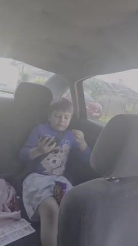 A young boy with brown hair sits in the backseat of a car, looking at his phone. He is wearing a blue long-sleeved shirt with a cartoon character on it and grey shorts. He has a seatbelt on. A bag of chips is on his lap. The car interior is visible, as is a window showing trees and a red car outside. The video is shot from the perspective of someone in the front seat.