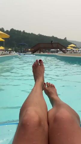 This is a first-person perspective video taken from a person's legs and feet submerged in a swimming pool. The person is lying on their back on an inflatable tube. The water is a clear turquoise color and has ripples. In the background, there is a wooden bridge, several people, lounge chairs, umbrellas, and trees. The sky is overcast. The person's toenails are painted red. The camera pans slightly to the right and left, showing more of the pool area and the people within it. There are also several inflatable rings visible near the edge of the pool.