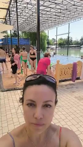 This is a selfie video taken at a crowded outdoor swimming pool area. In the foreground, a woman with dark hair and sunglasses on her head smiles and looks at the camera. She is wearing a swimsuit with a colorful patterned top and a pearl necklace. In the background, several people are visible enjoying the pool and the surrounding area.

The scene depicts a sunny day, likely in the afternoon, judging by the bright sunlight and the shadows cast. The setting appears to be a resort or a public pool with a large body of water. There are lounge chairs and umbrellas providing shade, as well as a wooden deck area around the pool. The architecture of the covered area suggests a modern design with a metal frame.

The activity taking place is leisure and recreation at a swimming pool. People are seen walking around the pool, some are in the water, and others are socializing. There's a sense of a lively and bustling atmosphere, with individuals of various ages, including children, present.

On the blue panel in the background, there is some visible text. It appears to be in a Cyrillic script, and parts of it are legible, such as "TAXOD" and a logo that resembles a bird or a winged symbol. The presence of Cyrillic text suggests the location might have historical ties to Russian influence or be in a region where this script is commonly used.

The overall mood conveyed by the woman in the foreground is relaxed and cheerful, suggesting she is enjoying her time at the pool. The presence of many people and the bright weather indicate a popular spot for recreation on a warm day.
