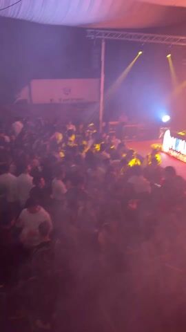 A crowd of people is gathered at what appears to be a nightclub or concert venue. A DJ booth with the name "MOONLIGHT" is on a stage, illuminated by various colored lights. The crowd is mostly composed of young adults, and they are standing, talking, and generally milling around. The venue has a tent-like roof, and there are lighting trusses overhead. To the left of the stage, there's a truck with "F. Torres" printed on it. There is smoke in the air, enhancing the light effects. A screen behind the DJ shows graphics. The lighting shifts from purple to orange and yellow.