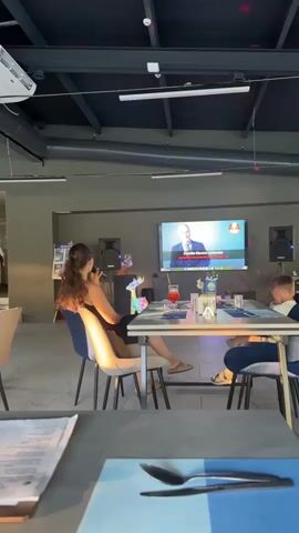 The video shows a karaoke session taking place in what appears to be a restaurant or bar. Two individuals, a woman with long brown hair and a young boy, are seated at a table facing a large screen displaying a karaoke video. The woman is holding a microphone and singing along, while the boy is also engaged with the screen.

The setting is indoors with a modern industrial aesthetic, featuring exposed ceiling beams, ductwork, and track lighting. Disco lights are projected onto the ceiling, creating a festive atmosphere. On the table, there are drinks, napkins, and cutlery. Two speakers are positioned on either side of the screen.

The screen displays a man in a suit singing, with Cyrillic text visible at the bottom. The text appears to be song lyrics. The overall mood seems relaxed and enjoyable, with the participants actively engaged in the karaoke activity. The lighting suggests it could be evening or a dimly lit indoor space. There is no visible indication of the weather outside. The location context provided is "Даниловка (алтайский), Russia".