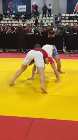 Two men are wrestling on a yellow and red mat. They are both barefoot and wearing shorts and rash guards. The man on the left is wearing a red rash guard and white shorts, and the man on the right is wearing a white rash guard and blue shorts. They are grappling and trying to take each other down. There are spectators watching in the background. The event appears to be a grappling competition or tournament. There are signs visible in the background, including one that says "RODA".