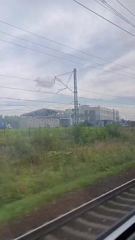 This is a video taken from a moving train looking out a window. The camera is angled slightly downwards and to the right, showing the train tracks in the foreground. To the left of the tracks, there is a grassy embankment with some scattered yellow flowers. Further in the distance, there is a long, multi-story building with a light gray facade and many windows. To the left of this building, there appears to be an industrial structure with a metal roof. The sky is overcast with scattered clouds. There are overhead power lines for the train visible throughout the video. The train is moving, causing a slight blur in the foreground and midground.