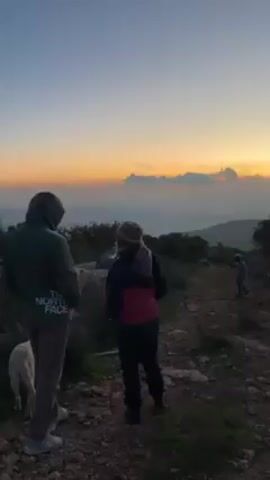 The video shows three people and a dog on a rocky path, silhouetted against a sunset. One person is wearing a green jacket with "THE NORTH FACE" printed on the back and khaki pants. The second person is wearing a hat, a dark jacket with a pink backpack, and dark pants. The third person is further down the path, a child, wearing dark clothes. The dog is a light color, standing near the first person. The sky is orange and pink near the horizon, fading to a light blue above. The landscape is hilly, with scattered trees and bushes.