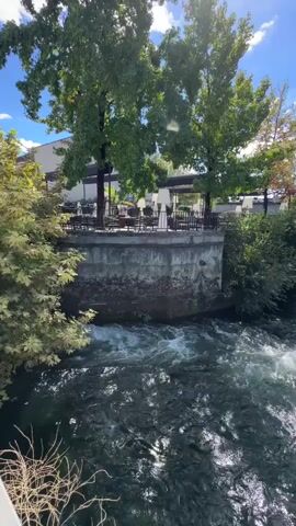 A fast-flowing river occupies the foreground, its surface disturbed by turbulent white water. Beyond the river, a raised concrete wall supports a paved area with trees and outdoor seating. A building with large windows is visible behind the seating area. Further in the distance, a concrete bridge spans the river, with cars crossing it. The sky is blue with scattered white clouds. The overall scene depicts a riverfront area with a restaurant and a bridge on a clear day.
