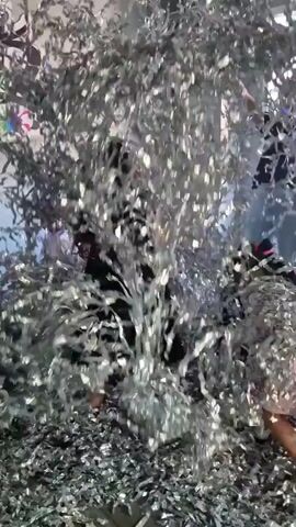 A group of people are inside a room with silver streamers falling all around them. In the foreground, a person in a black dress is visible on the left, partially obscured by streamers. To their right, a person with dark hair, also in a black outfit, is visible. Further back and to the right, another person wearing a pink garment is standing and looking towards the left. More individuals are present in the background, their features less distinct due to the dense covering of streamers. The room appears to be an indoor space, possibly an office or studio, with shelves and a window visible in the background. The streamers are creating a chaotic and celebratory atmosphere.