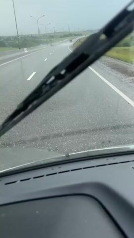 This media file captures a point-of-view shot from inside a vehicle driving on a wet highway during what appears to be daytime. The perspective is looking through the windshield, which is streaked with rain and has windshield wipers visible in the foreground. The highway stretches ahead, with white lane markings on a gray asphalt surface. The road is wet and reflects the overcast sky, suggesting rain or recent rainfall. The surrounding landscape consists of green fields and trees on either side of the road, with utility poles and distant structures visible. The sky is a uniform gray, indicating cloudy or stormy weather. There are other vehicles on the road in the distance. The overall mood is one of driving through inclement weather. No visible text is present in the media file.