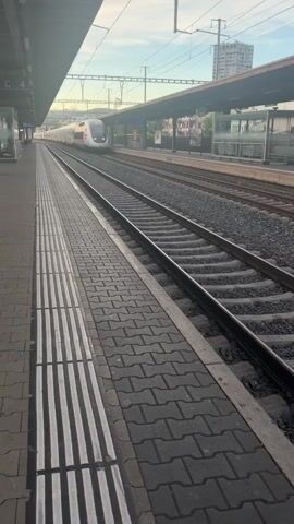 The video shows a train at a station. The train is white and red and appears to be a high-speed train. It is moving along the tracks. The station platform is made of gray pavers and has a tactile paving strip along the edge. There are overhead wires and a building in the background. The sky is cloudy. The video appears to be filmed from the perspective of someone standing on the platform.