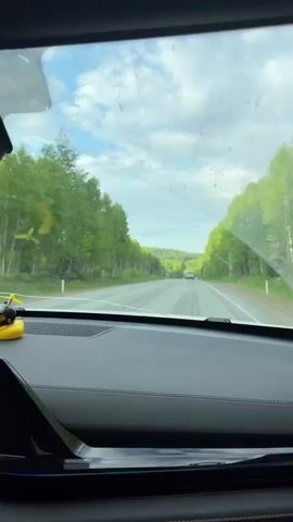 The camera is positioned inside a car, filming the road ahead through the windshield. The car is driving on a highway lined with trees on both sides. The sky is partly cloudy. On the dashboard, there is a small yellow rubber duck wearing a helmet and goggles. The car's infotainment screen displays a map and media player interface. The car is playing music. A rectangular air freshener hangs from the rearview mirror.