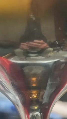 The video captures a close-up view of a copper coil wrapped around a black object, likely a component of some device. The coil sits on a metallic surface, which is partially obscured by what appears to be a clear, reflective material, possibly glass or plastic. This material has red accents visible along its edges, suggesting it might be part of a container or an external casing. Below this surface, a yellowish, possibly liquid substance is visible, reflecting light and creating a shimmering effect. The overall environment is dimly lit, with a focus on the intricate details of the coil and its mounting.