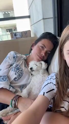 The video shows a woman holding a small white dog while sitting next to a young girl. The woman is wearing a white blouse with blue patterns and several bracelets on her left wrist. The dog is nestled in her arms, with its eyes closed. The girl is wearing a striped shirt and has her right hand raised to her face. Both the woman and the girl are looking towards the camera. They appear to be indoors, possibly on a balcony, with a building visible in the background. The lighting suggests it is daytime. The woman is smiling gently, while the girl has a more neutral expression. There is a box visible on a shelf in the background.