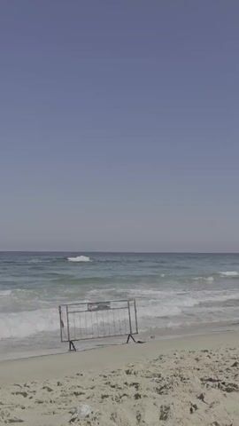 A static shot of a beach with a metal barrier in the foreground. The barrier is on the sand, and waves are breaking on the shore behind it. The sky is clear and blue.
