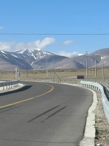 The video captures a wide shot of a winding paved road in a mountainous, arid landscape, likely Gyirong County, China. The camera pans slowly from left to right, revealing the scenic environment.

In the foreground, a dark gray asphalt road curves gently to the right, marked with a continuous yellow line. A metal guardrail with white posts runs along the right edge of the road, separating it from the drier, brownish terrain. Long shadows, presumably from utility poles, stretch across the road.

In the midground and background, numerous tall utility poles with power lines extend across the landscape, paralleling the road. Several speed limit signs are visible, indicating limits of 30 km/h and 50 km/h. A small blue sign with unreadable white text is also present on the right side.

Majestic mountains dominate the background, with their peaks partially covered in snow or ice. The sky is a clear, bright blue with scattered white clouds, suggesting a sunny day. The overall scene depicts a remote, high-altitude road amidst rugged natural beauty. No people or vehicles are visible.