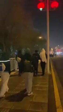 The video shows a group of people walking along a sidewalk in an urban setting at night. The sidewalk is lined with trees and illuminated by streetlights and red lanterns. A road runs parallel to the sidewalk, with cars driving by. Buildings are visible in the background, illuminated with lights. The people are walking in the same direction, away from the camera. Some are wearing casual clothing, while others have backpacks. The overall atmosphere is dark and slightly hazy, with a sense of movement and activity in the city.