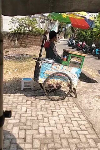 A vendor operates a food cart in an outdoor setting. The cart has a colorful umbrella and displays text that appears to be in Indonesian. Two young people, possibly students based on their uniforms, are interacting near the cart. One is seen holding a device or tool, while the other is looking on. The ground is paved with bricks, and there are buildings and trees in the background. The scene suggests a daytime activity, possibly a street market or a school event.