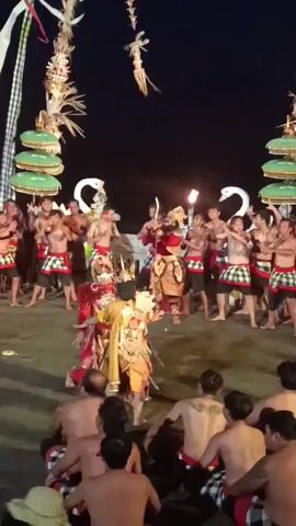 The video shows an outdoor night performance. In the center, two performers, one in a red and gold costume and another in a yellow and gold costume, are engaged in a dance. A group of men, shirtless and wearing checkered sarongs, surround them. Some hold torches. In the background, there are tall, decorated structures with tiered, green umbrellas. The ground appears to be sand or dirt. The lighting is dim, with spotlights illuminating the performers and torches providing additional light. The audience is visible in the foreground, seated and facing the performance.
