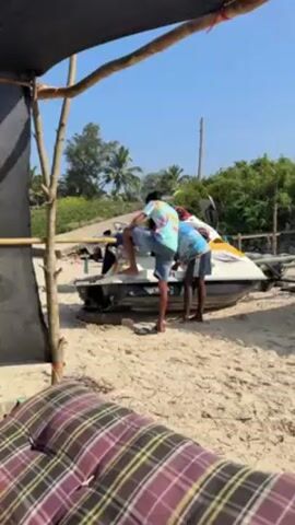 From under a makeshift awning on a sunny beach, two men are focused on a white and yellow jet ski resting on a sandy surface. One man, wearing a tie-dye shirt and blue shorts, has his leg on the jet ski. The other man, dressed in a similar tie-dye shirt and dark shorts, bends over towards the watercraft.  A third man in a white shirt and baseball cap watches from nearby. Lush greenery is visible in the background under a clear blue sky.