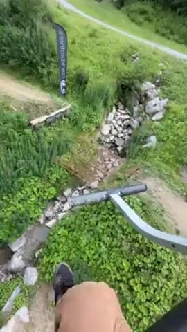 The video shows a person riding a ski lift, possibly in the summer, as the ground below is covered in green grass and trees. A dirt path winds its way up the mountain, and there are occasional flag markers along the path. The sky is overcast, with patches of blue visible. The view is from the perspective of the rider, looking down and ahead. The seat and footrest of the lift are visible in the foreground.