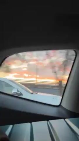 The video shows a view from inside a car, looking out the rear passenger side window. Another white car is visible driving alongside the videographer's car.  The sky is visible through the car window and shows a sunset with orange and pink hues.  Trees and street lights are visible on the roadside.  The videographer's car appears to be in motion.