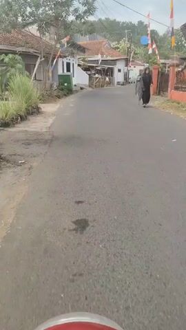 The video shows a view from a moving vehicle, likely a motorcycle, traveling down a paved road in a rural or suburban area. The road curves to the right. On the left side of the road are houses with tile roofs and some vegetation. On the right side of the road, there is a fence and more houses. Indonesian flags, red and white, are visible flying from poles along the road, suggesting a national holiday or celebration. Two people wearing dark clothing and headscarves are walking away from the camera on the right side of the road. The sky is overcast.