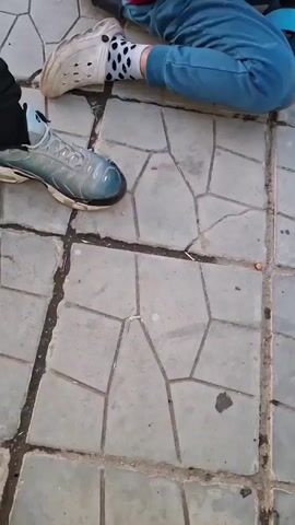 The video opens with a young child sitting on grey paved ground, holding onto the handlebars of a blue scooter. The child is wearing a red vest, blue pants, and white socks with black polka dots, paired with white clogs or sandals. Part of the text "THE FUT" is visible on the red vest. Another person's foot in blue and black athletic shoes is in the foreground. The scene then shifts to a young male walking outdoors, holding a white packet or carton. He wears a black jacket with a lime green upper arm detail. The background shows a paved road, houses with red tiled roofs, and green foliage. The video concludes with a shot of the same young male, now facing the camera, wearing a black Adidas hoodie with white stripes on the sleeves. The background features residential houses and power lines under a bright, early evening sky in what appears to be a residential area in Harlets, Bulgaria.