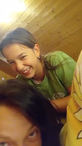 The video shows two people in close proximity, with one person, presumably a child, leaning over the other. The child is wearing a green t-shirt and has long, dark hair pulled back in a ponytail. They are smiling and appear to be tickling or playing with the hair of the person in the foreground. The person in the foreground has dark hair and is looking directly at the camera with a neutral expression. The background consists of a wooden paneled wall and ceiling, suggesting an indoor setting, possibly a room with a sloped ceiling like an attic or loft. The lighting is warm and appears to be artificial. There is a pillow with a yellow and white pattern visible on the right side of the frame. The overall impression is of a playful and intimate moment between two individuals.
