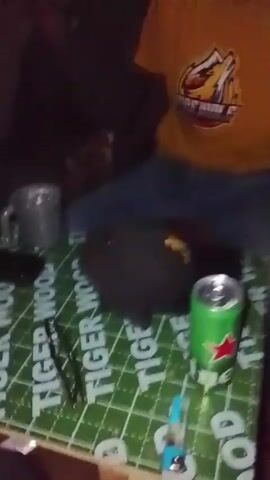 The video captures a close-up, top-down view of a table in a dimly lit indoor setting, likely in Durumi, Nigeria, during the evening or night. The table is covered with a green and white checkered mat or cloth, prominently featuring the repeated text "TIGER WOOD."

Various items are scattered on the table, suggesting a social gathering. These include a brown glass bottle, a green Heineken beer can, a dark bottle with a blue cap, and a silver metallic mug. A pair of black-framed glasses and a small white packet are also visible.

Parts of at least two individuals are seen around the table. One person is wearing blue pants and a brown or yellow top, while another wears dark pants and white shoes. The camera appears to be held by someone seated at the table, capturing the casual atmosphere of people gathering, possibly drinking and socializing.