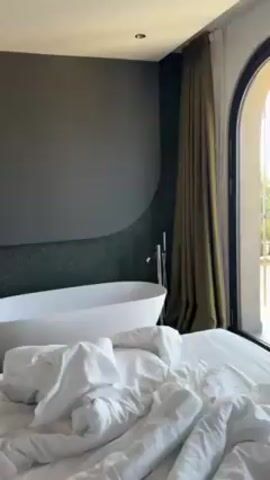 The video shows a bedroom, with an unmade bed in the foreground. In the corner of the room is a modern freestanding tub. To the right of the bed, an arched glass door opens to a balcony. The camera pans towards the balcony, showing a view of a lake with surrounding buildings and trees on a sunny day. The balcony has metal railings and a couple of chairs.