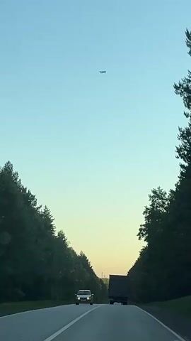 The video captures a forward-moving perspective from a vehicle traveling on a two-lane road in a rural or forested area. Dense green trees line both sides of the road, creating a natural corridor, with the foliage appearing rich and dark. Ahead, a silver sedan is visible in the left lane, and a large, dark truck is seen further down the road in the right lane. The road surface has clear white markings, including a solid line on the right shoulder and a broken line dividing the lanes.

The sky above is clear and transitions from a light, pale blue at the top to a warm, soft yellow or golden hue closer to the horizon, suggesting either early morning or late evening light. High in the sky, a small aircraft or airplane is visible, silhouetted against the clear sky, flying horizontally in the distance. The overall scene conveys a sense of travel through a peaceful, natural landscape under a serene sky. The location context provided is Iiul'skoe, Russia.