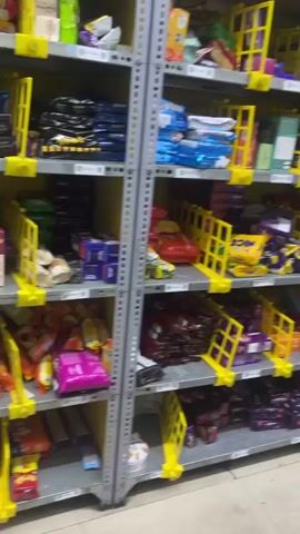 The video opens with a view of a storage area or retail space, featuring tall metal shelving units filled with various packaged goods, possibly snacks or small consumer items. The shelves are organized with numerous yellow plastic bins or dividers, holding different products.

The camera then pans to a young man sitting on a black office chair beside a desk. He is wearing a grey t-shirt with a yellow design and black track pants. He is engrossed in his smartphone, holding it horizontally with both hands, suggesting he is playing a game or watching a video. He appears focused and relaxed.

On the desk next to him, there is a dark-colored printer or scanner and another mobile phone. In the background, the shelves continue, extending along the wall and filled with more inventory. The overall setting resembles a stockroom, a small convenience store, or a delivery preparation area, likely indoors in Palghar, India. The lighting is artificial, suggesting it could be any time during operating hours. Some faint text is visible on a yellow sign on the wall behind the man, but it is illegible. Another label on the desk next to the printer reads "PERSONAL" and "RS O," though it is partially cut off.