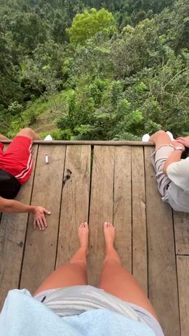 The video opens with a low-angle shot of three people sitting on a wooden platform, their legs and feet dangling over the edge. The person recording is barefoot. A person in red shorts sits to the left, and a person in gray shorts sits to the right. Between them, on the wooden planks, is a small, white object that resembles a cigarette lighter. The camera then tilts upwards to reveal a vast, lush green forest extending into the distance. Mountains are visible in the background. The sky is cloudy, with patches of sunlight breaking through. The overall scene suggests a scenic viewpoint or observation deck overlooking a forest and mountain range.