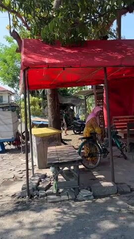 The video shows an outdoor scene under a red tent. A woman wearing a yellow dress and a pink headscarf is standing next to a bicycle. She appears to be interacting with the bicycle, possibly preparing to ride it. In the background, there is a cart with white objects on it, a motorcycle, and trees. The ground is paved with stones and dirt. The sun is shining, casting shadows.