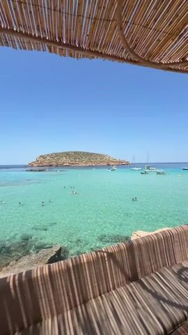 This is a video capturing a serene beach scene, likely in Sant Josep de sa Talaia, Spain, as suggested by the location context. The video opens with a view from under a woven bamboo canopy, offering shade and a sense of relaxed ambiance. Beyond the canopy, the crystal-clear turquoise waters of the Mediterranean Sea are visible, teeming with life and activity.

The main subjects are the beautiful natural scenery and the people enjoying it. The water is the most striking element, with its vibrant aquamarine hue and translucent quality, revealing the sandy bottom in some areas. A small, rocky island, covered in sparse green vegetation, sits in the distance, adding to the picturesque landscape.

Scattered across the water are numerous sailboats and smaller vessels, suggesting a popular spot for boating and leisure. Closer to the shore, several people are swimming and enjoying the water, their figures appearing as small silhouettes against the bright expanse. The overall atmosphere is one of tranquility and vacation enjoyment.

The time of day appears to be midday or early afternoon, indicated by the bright sunlight casting distinct shadows from the canopy onto the seating area. The weather is clearly sunny and fair, with a clear blue sky dominating the upper portion of the frame.

Notable details include the texture of the woven canopy and the cushioned seating area, which suggest a comfortable, perhaps resort-like, setting. The gentle ripples on the water indicate a calm sea. There is no visible text in the media file. The emotions evoked are those of peace, relaxation, and the joy of a perfect summer day at the beach.