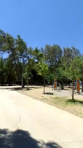 The video shows a park area on a sunny day with a clear blue sky. There are several trees, some with green leaves and others with sparser foliage. In the foreground, a paved path curves through the scene. Scattered throughout the park are various outdoor exercise equipment stations, mostly featuring yellow and orange components. A person is visible near one of the exercise stations, appearing to be engaged in some activity. Further into the park, there is a wooden gazebo or pergola structure with benches underneath. The ground is covered in dry grass and dirt. The camera pans slowly across the scene, offering a broad view of the park's amenities and natural elements.