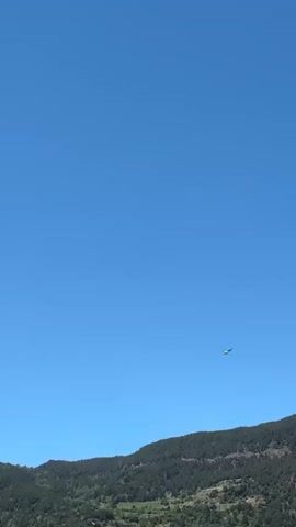 A helicopter, partially yellow and dark-colored, flies across a clear blue sky from right to left. Below, a forested mountain range stretches across the horizon, covered in dense green trees. The scene depicts a bright, sunny day with no clouds visible. The location is set in Encamp, Andorra, with mountainous terrain characteristic of the region. No people or visible text are present in the video.