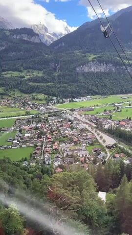 The video shows an aerial view of a small town nestled in a valley between lush green mountains. A cable car or gondola wire is visible in the foreground, indicating the viewpoint is from a moving vehicle. The town consists of houses with red-tiled roofs and appears to be situated alongside a winding road. Green fields surround the residential areas, and a river or stream runs through the valley. The mountains are heavily forested, with some rocky peaks visible in the distance under a partly cloudy sky. The overall scene depicts a serene, mountainous landscape with a village at its base.