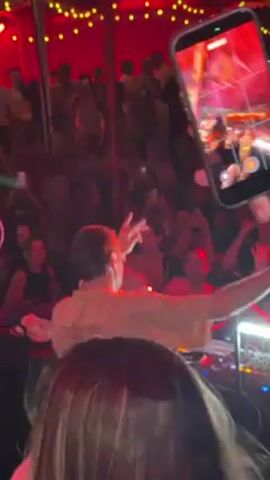 The video captures a lively club scene from the perspective of someone in the crowd, who is holding up a smartphone to record. The focus is on a DJ, who stands behind a mixing console and interacts with the energized crowd. The camera person wears a red wristband and is jostled by other patrons, the device moving up and down. The club is dimly lit with red lighting, and the crowd is dense. Many people are visible behind the DJ. String lights twinkle overhead. The video offers a close-up view of the DJ's actions and the electric atmosphere of the club.