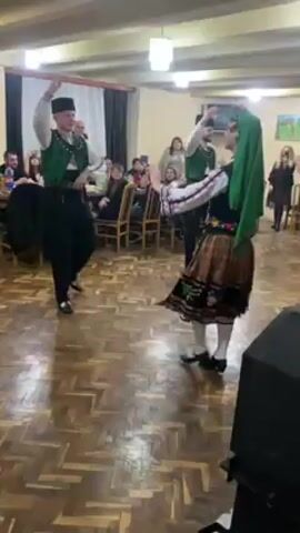 The video shows a man and a woman dancing in what appears to be a ballroom or event space. They are both wearing traditional folk costumes with intricate embroidery. The man holds a wooden stick in his hand. The floor is made of wooden planks arranged in a herringbone pattern. Several people sit at tables in the background, watching and filming the dancers with their phones. There is a speaker on the floor to the right of the dancers. The room is brightly lit by ceiling lights.
