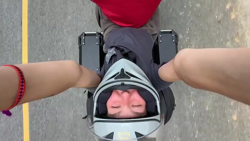 The video features a motorcycle journey through a mountainous, green landscape.

The first shot (0:00-0:01) is a first-person view from a passenger looking down at the motorcycle rider. The rider wears a light grey full-face helmet with a clear visor, a dark grey t-shirt, and possibly beige shorts. Their eyes are closed, potentially due to wind or sun. Two black rectangular storage boxes (panniers) are visible on either side of the motorcycle. The passenger's legs are visible, one wearing a red and black bracelet. The motorcycle is moving on a paved road with a yellow line. The visible text on the helmet chin bar is "ROYAL".

The subsequent shots (0:01-0:03) are filmed from behind the rider, looking forward. The rider is now wearing a black and white full-face helmet with a dark visor and complex graphics, a bright red puffy jacket, and appears to have long hair tied back. The motorcycle travels on a paved road with a yellow line, bordered by lush green foliage and trees. The background features rolling green mountains under a partly cloudy sky. A small wooden structure is visible on a hillside, and an area of disturbed earth with an excavator suggests recent construction or a landslide. The prominent visible text on the back of the helmet is "YOHE". The overall scene depicts a scenic road trip through a verdant, hilly region.
