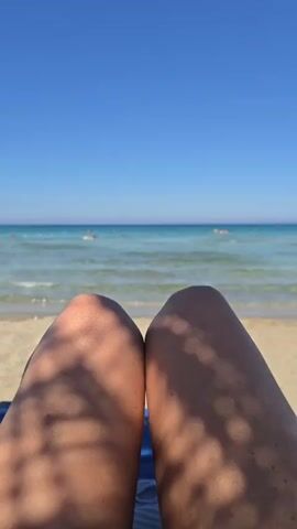 The video shows a first-person perspective of someone lying on a beach. The viewer sees the person's legs from the knees down, with shadow patterns from palm fronds falling across them. Beyond the legs, a sandy beach slopes down to a calm, turquoise ocean. In the distance, the ocean meets a clear blue sky. Several people can be seen in the water, some swimming and others appearing to float. The sun is shining brightly, casting distinct shadows. The scene evokes a feeling of relaxation and a summer vacation.