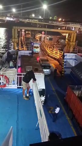 This is a night-time aerial view from a ferry showing vehicles boarding. A man in a black jacket and shorts is leaning over a railing on the ferry's deck. Below him, on the blue deck of the ferry, are several trucks and at least one car. Two individuals, one in a reflective orange vest and another in a dark uniform, are standing near the vehicles. The ferry is docked, and a ramp is extended from the shore to the ferry, with more trucks visible on the ramp and beyond. The water is visible to the left of the ferry. Lighting from streetlights and the ferry illuminates the scene.