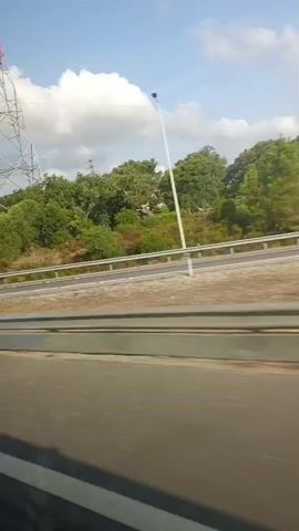 The video shows a view from a vehicle traveling on a road. On the left side of the road, there is a large metal tower with red and white markings, and further down, a shorter tower with antennas. There is a bright yellow sound barrier along the side of the road. Green trees are visible behind the barrier. The sky is blue with scattered white clouds. A yellow arrow sign pointing left is also visible on the left side of the road. The road appears to be a highway with multiple lanes and white lane markings. The footage is recorded from a moving vehicle, indicated by the blurring of the scenery.