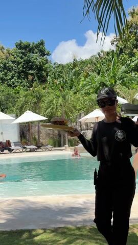 A woman in sunglasses, a black t-shirt, and a black baseball cap stands outdoors next to a swimming pool. She is holding a yellow tray with a large sandwich, a small wire basket of french fries, and a small black bowl of ketchup. Behind her is a swimming pool, several white lounge chairs, white umbrellas, and trees. The woman appears to be a waitress, bringing the food to a customer. The camera pans around the tray of food once it is set down, highlighting the details.