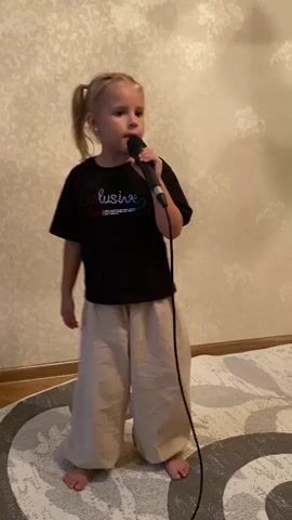 A young girl with blonde hair in pigtails stands in front of a textured beige wall, singing into a microphone. She is wearing a black t-shirt with the word "Exclusive" on it and loose, light-colored pants. She is barefoot and standing on a rug with a circular pattern. The microphone is connected to a wire that trails down and out of frame. The girl's mouth is open as she sings. The lighting is indoor and appears to be from above.