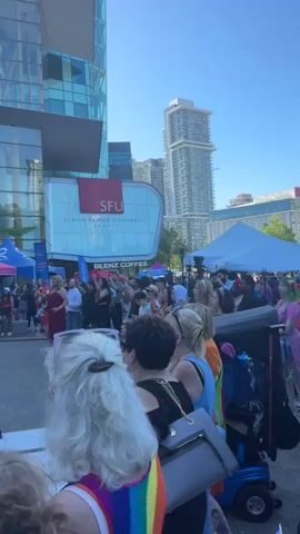 A video shows a daytime outdoor event. In the foreground, the backs of several people are visible, some with blonde hair. Behind them, a crowd gathers, with several tents set up, one pink and blue. A large building with a glass facade and an SFU (Simon Fraser University) sign is in the background. Further back, tall skyscrapers are visible.

The video pans to a stage where a person in a pink outfit and silver boots appears to be performing. Another person in a white top and shorts is interacting with the performer on stage. A banner above the stage reads "Surrey Pride.ca" with a rainbow flag. A sign for "Prospera Credit Union" is visible on the stage. The overall atmosphere seems festive.