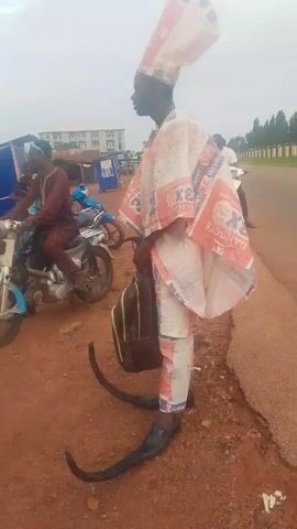 A man stands on the side of a road wearing an outfit constructed from bags labeled "Dangote Cement." He has a tall hat, a robe, and pants made from the same material. His shoes are black with long, curved points. He carries a bag, possibly a backpack, over his shoulder. The road is paved and has some traffic, including motorcycles. The shoulder of the road where the man stands is red dirt. In the background, there are buildings, trees, and other people. The sky is overcast.